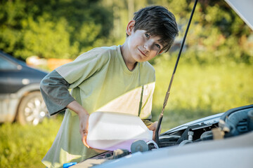 A young boy pours windshield washer fluid into a car's engine compartment on a sunny day. The scene is set outdoors with a blurred green background.