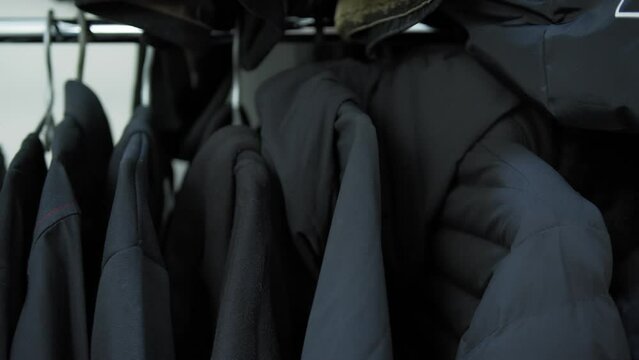 Camera slides past close-up wardrobe with clothing on hangers: sweaters, hoodies, jackets, coats, of same type, dark gray black colors, on light background. Monochrome and monotony clothes concept .