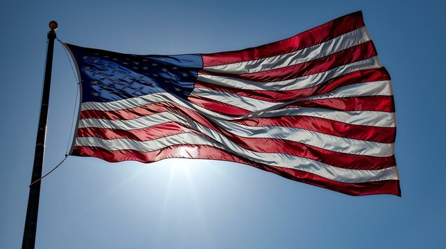The image shows a large American flag waving in the wind against a clear blue sky. The flag is backlit by the sun, which creates a dramatic effect. - Powered by Adobe