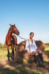 A young girl in a white dress sits on the dry grass next to the horse.