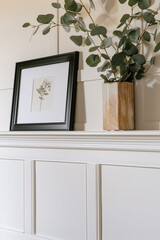 Close-up of a horizontal poster in a black frame standing on a shelf leaning against white wall panels next to eucalyptus branches in a beige vase. Natural lighting