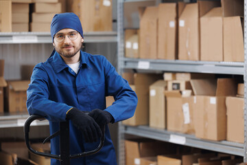Man loader in uniform holding trolley with product in warehouse. Service of factory logistic import...