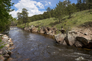 Beautiful view of a mountain creek with large boulders and rocks against a green tree filled background and partially cloudy blue skies.