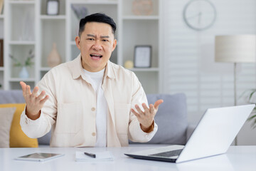 Man expressing frustration during a video call meeting at home. He is seated at a desk with a laptop and paperwork.