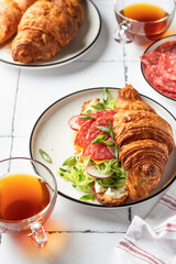 Croissant sandwich with salami sausage, cheese and cucumber in a plate with cup of tea for tasty breakfast on white tile table