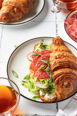 Croissant sandwich with salami sausage, cheese and cucumber in a plate close up with cup of tea for tasty breakfast on white tile table