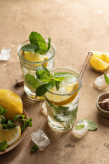 Two glasses of mojito cocktail or tonic with lemon, mint and ice on brown textured background. Refreshing summer drink, text space