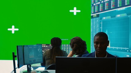 Diverse group of business specialists solve their daily tasks in control room with big greenscreen,...