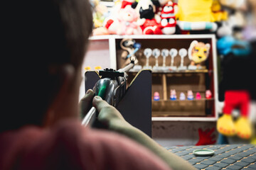 A young boy aiming a rifle at a target in a shooting game booth. The booth is filled with plush...