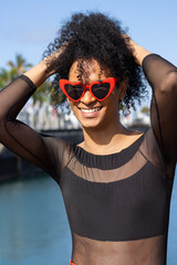 Non-binary person with afro hair and heart-shaped sunglasses, holding their hair while smiling