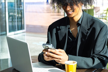 Non-binary executive person wearing a black suit and afro hair, smiling while typing on their mobile phone. Laptop and coffee cup