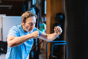 A senior man performs martial arts by punching a bag in a gym.