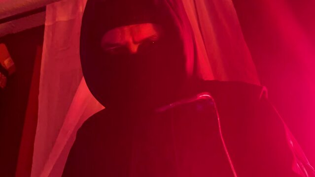 A robber burglar is breaking into a home with a black mask and weapon with red light shining on the curtain in the night darkness for a home surveillance or security concept. 