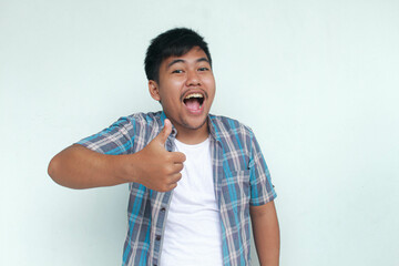 Excited and happy Asian man gives thumbs up hand gesture of approval, isolated by gray background.