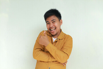 Excited and happy Asian man gives thumbs up hand gesture of approval, isolated by gray background.