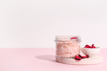 Jar of natural body scrub and flower petals on color table against light background