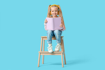 Cute little girl in headphones listening to audiobook with book while sitting on stool against blue background