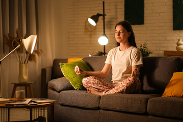 Young woman in pajamas meditating on cozy sofa in living room at night