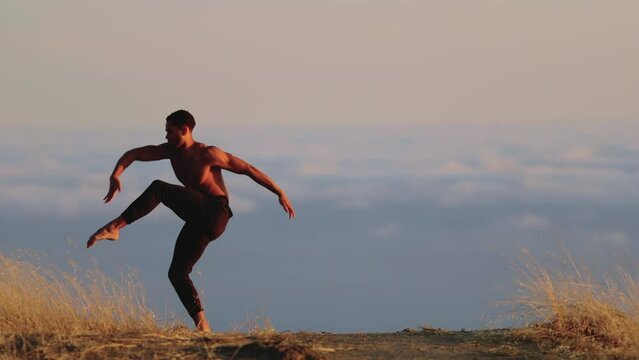 Male ballet dancer doing a choreographic dance move standing on a rock at sunset. Heavenly clouds are in the background