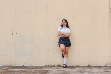 Fototapeta premium Young asian woman stands confidently against an aged wall outdoors, showcasing casual fashion and a relaxed attitude.