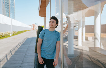 Young man standing outdoors with a yoga mat on his shoulder, leaning against a glass wall, wearing blue t-shirt. Modern urban background with buildings and clear sky. Relaxed and thoughtful expression