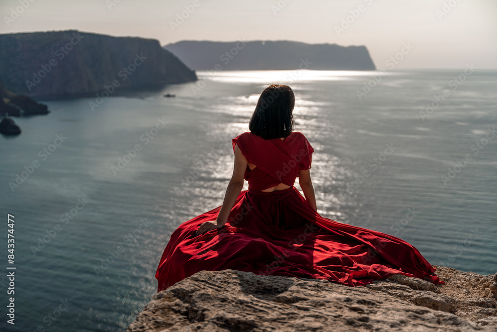 Wall mural A woman in a red dress is sitting on a rock overlooking the ocean. The scene is serene and peaceful, with the woman taking in the beauty of the water and the sky.