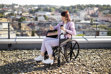 Confident woman in wheelchair holds cup of coffee while using smartphone on sunny rooftop. Captures independence, urban lifestyle, and modern technology.