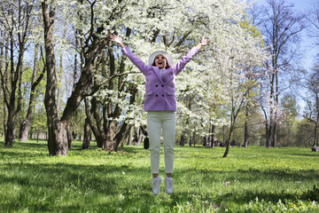 Emotional, joyful bouncing woman in white pants, lilac jacket, and white hat bouncing hard against a blooming tree in a spring park.