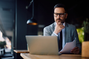 Pensive entrepreneur using laptop while analyzing business reports in  café.