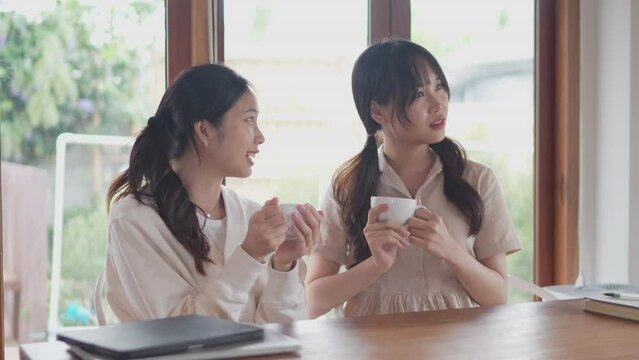 Two Asian female friends are chatting at a table and drinking hot coffee together at home