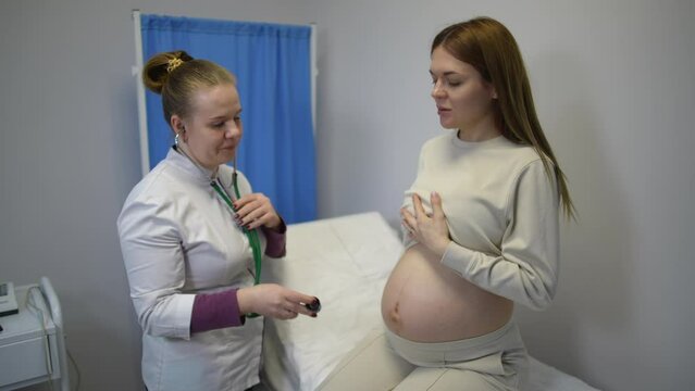Slow motion. A female doctor puts on a stethoscope and applies a diaphragm to the belly of a pregnant woman, sitting on an examination couch in a doctor's office during a medical examination at a