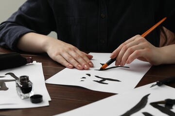 Calligraphy. Woman with brush and inkwell writing hieroglyphs on paper at wooden table, closeup