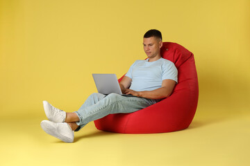 Handsome man with laptop on red bean bag chair against yellow background