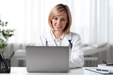 Naklejka premium Smiling doctor with laptop having online consultation at table in office