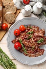 Slices of tasty fried bacon with rosemary and tomatoes served on wooden table, flat lay