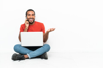 Young Ecuadorian man with a laptop sitting on the floor isolated on white background keeping a conversation with the mobile phone with someone
