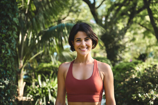Happy smiling fit sporty active slim confident young Hispanic woman wearing sportswear top enjoying summer sunny day looking at camera standing in green nature park or forest. Portrait.