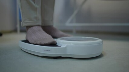 Close-up of human legs in pants and socks while being weighed on a bathroom scale that sits on the floor in a clinic