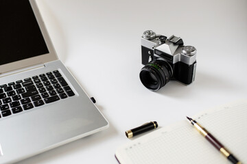 A view of the lens of an old vintage camera, which stands on a white table next to a gray laptop, and a notepad for recordings.