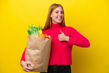 Young redhead woman holding a grocery shopping bag isolated on yellow background giving a thumbs up gesture