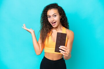 Young Arab woman isolated on blue background taking a chocolate tablet and surprised