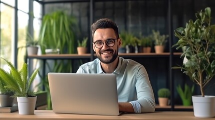 A man with glasses smiles at the camera while working on his laptop in an office setting
