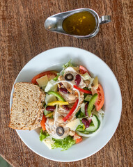 Top view of vegetable salad with olives, cheese and slice of bread on a white plate and dressing on the side