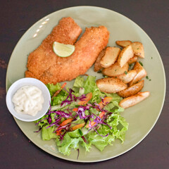 Top view of breaded fish fillet served with vegetable salad, potato wedges and tartar sauce
