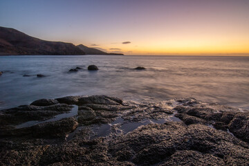 View of the sunrise over the Atlantic Ocean. View over a stone beach of a volcanic island. Cold lava stones by the sea on the beach at Cueva de Playa en Tarajalejo, Fuerteventura, Canary Islands, Spai