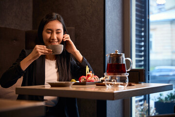 Positive Asian woman drinking tea in japanese restaurant and talking phone