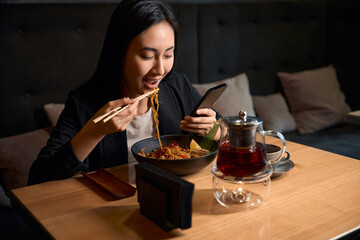 Hungry young woman eating noodles reading message on smartphone