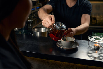 Courteous barman pouring fresh tea into the cup of visitor