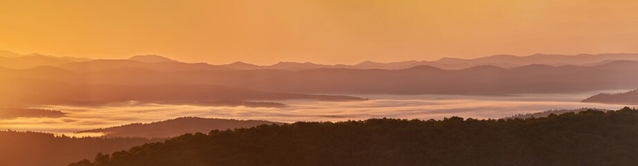 A panorama of a mountain valley in the morning mist. Picturesque dawn in the summer mountains. Warm sunlight