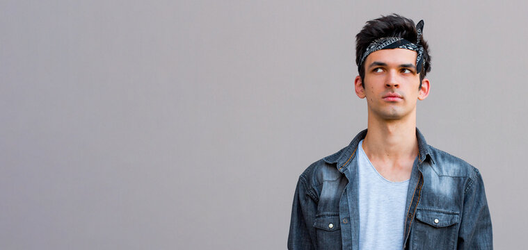 Young attractive male with skeptic look, raises eyebrow in bewilderment, has some doubts and uncertainty, looks curiously aside, isolated over gray background